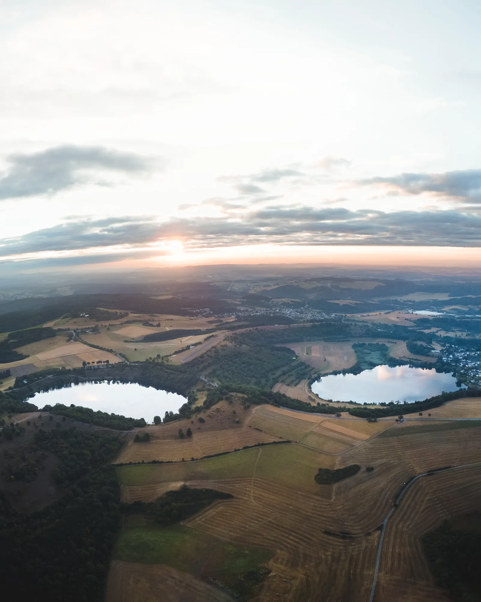 Aerial view at sunrise of two round crater lakes, the Eifel maars, surrounded by fields and forest